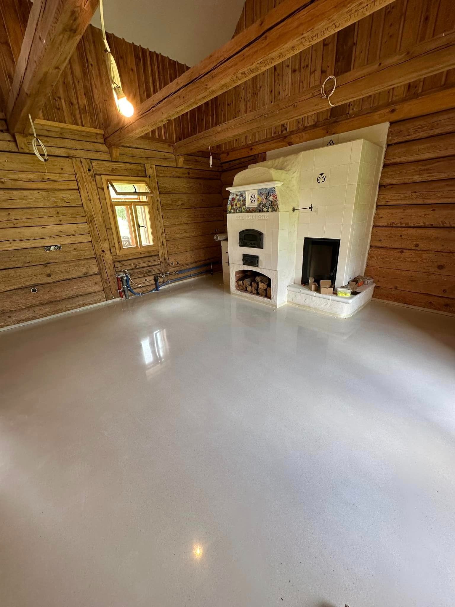 Jasnoszara, gładka podłoga w drewnianym wnętrzu z piecem kaflowym. Widoczne instalacje przy ścianie, surowy wystrój. Podłoga o wyglądzie lastryko.
