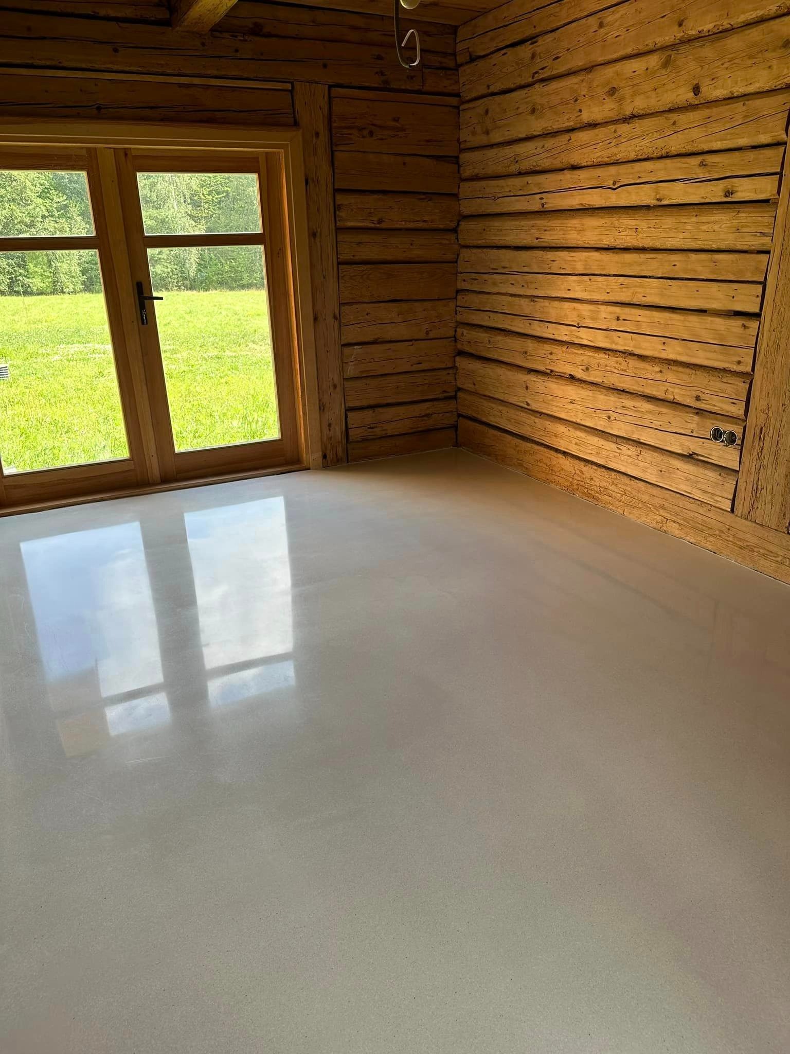 Jasna, gładka posadzka w rustykalnym wnętrzu z drewnianymi ścianami i oknem z widokiem na zieleń w Krakowie. Powierzchnia odbija światło okienne.