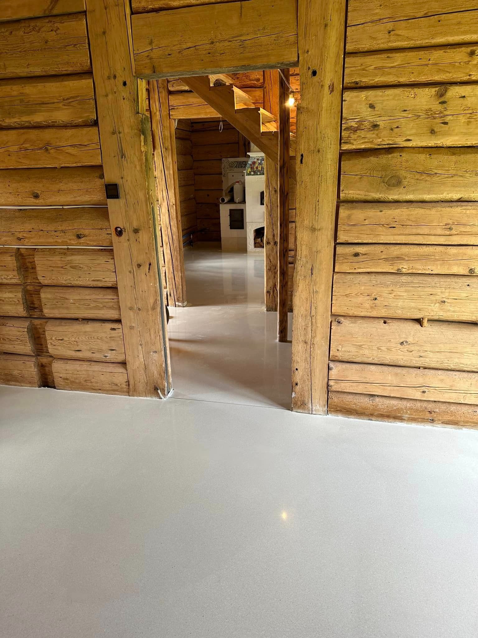 Jasnoszara, gładka posadzka żywiczna w drewnianym domu z bali. Widok na wnętrze przez drewniane framugi, schody i kominek w tle. Nowoczesne wykończenie w rustykalnym otoczeniu.