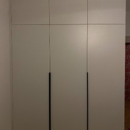 Volodymyr Khomliak - Biała szafa trzydrzwiowa z czarnymi, minimalistycznymi uchwytami, zabudowana od podłogi do sufitu, z widoczną podłogą z paneli.