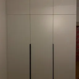 Biała szafa trzydrzwiowa z czarnymi, minimalistycznymi uchwytami, zabudowana od podłogi do sufitu, z widoczną podłogą z paneli.