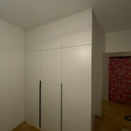 Volodymyr Khomliak - Biała szafa wnękowa z minimalistycznymi, pionowymi uchwytami na tle parkietu i widocznym fragmentem wzorzystej zasłony w głębi.