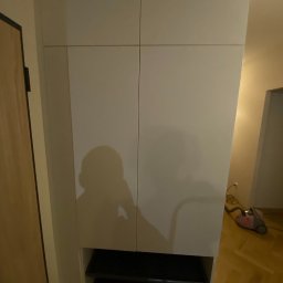 Volodymyr Khomliak - Biała szafa z frontami bez uchwytów i otwartą półką na dole, cień osoby widoczny na froncie, odkurzacz w tle.