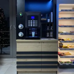 Automatyczny ekspres do kawy Saeco w zabudowie z szafkami w kolorze drewna i czarnymi pasami, obok regał z przekąskami, całość oświetlona niebieskim światłem LED.