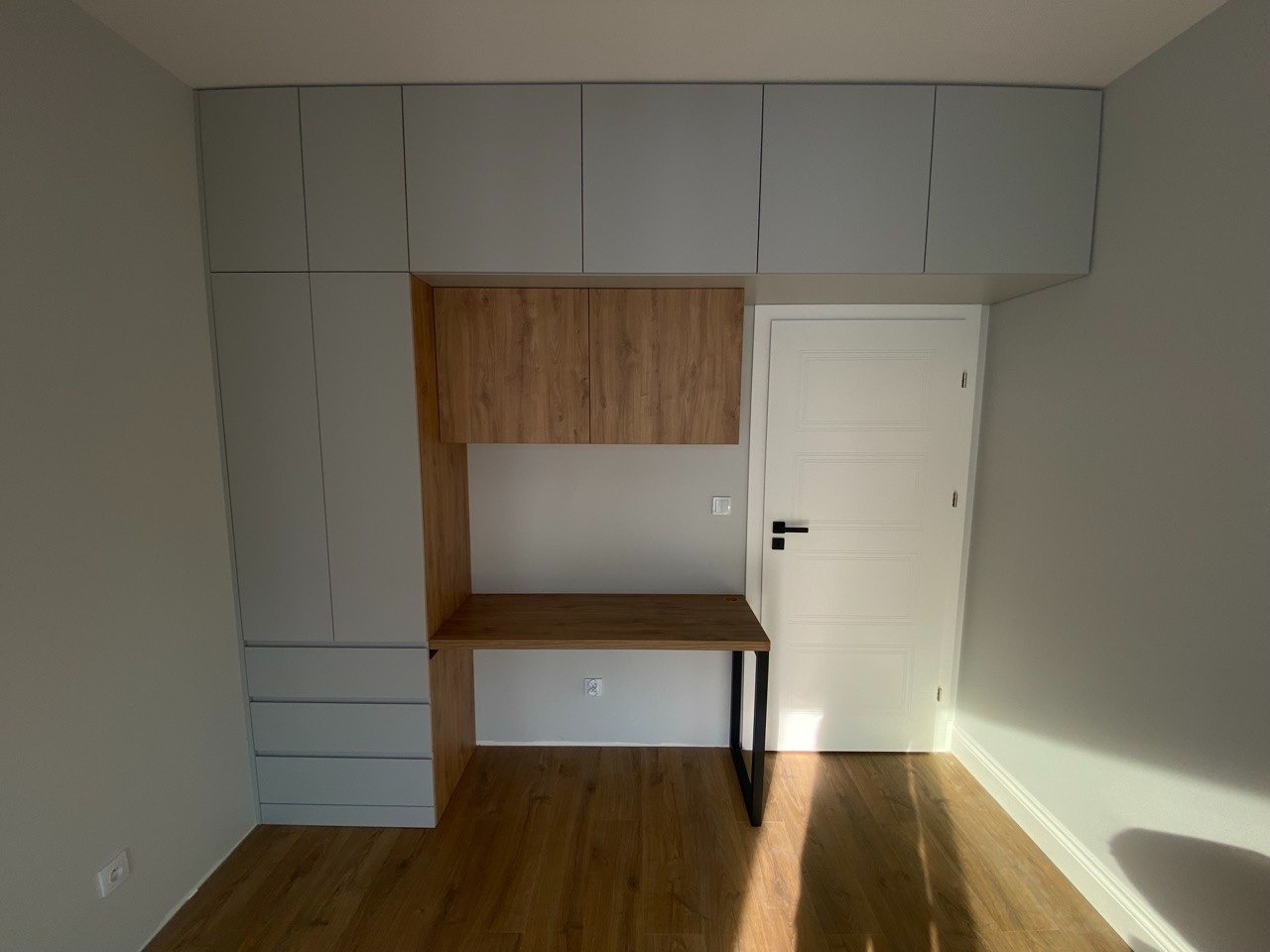 Szara zabudowa meblowa z drewnianym biurkiem i szafkami wiszącymi, dopasowana do wysokości pomieszczenia, zintegrowana z drzwiami.