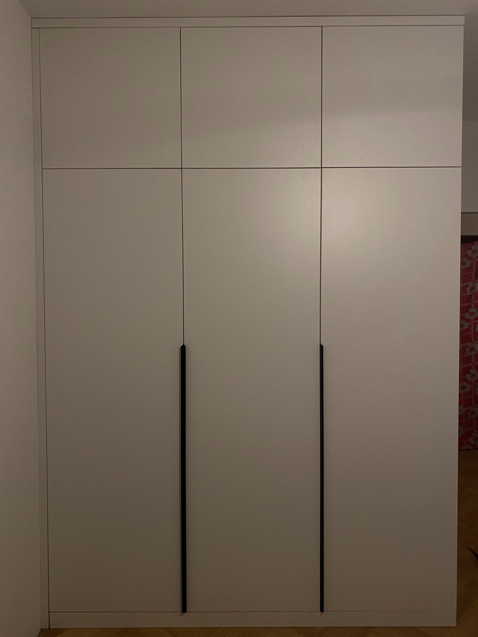 Biała szafa trzydrzwiowa z czarnymi, minimalistycznymi uchwytami, zabudowana od podłogi do sufitu, z widoczną podłogą z paneli.