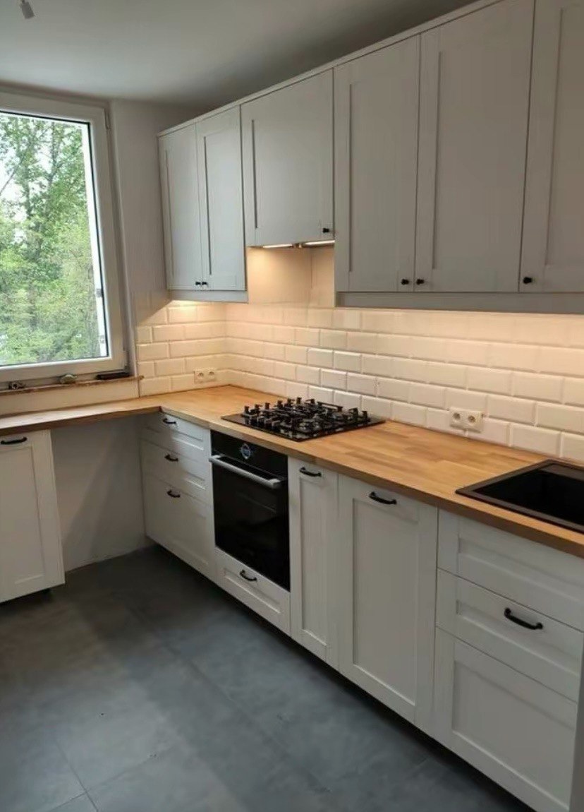 Jasna kuchnia z białymi szafkami z frezowanymi frontami, drewnianym blatem, białymi płytkami metra i czarnym zlewem oraz płytą gazową.