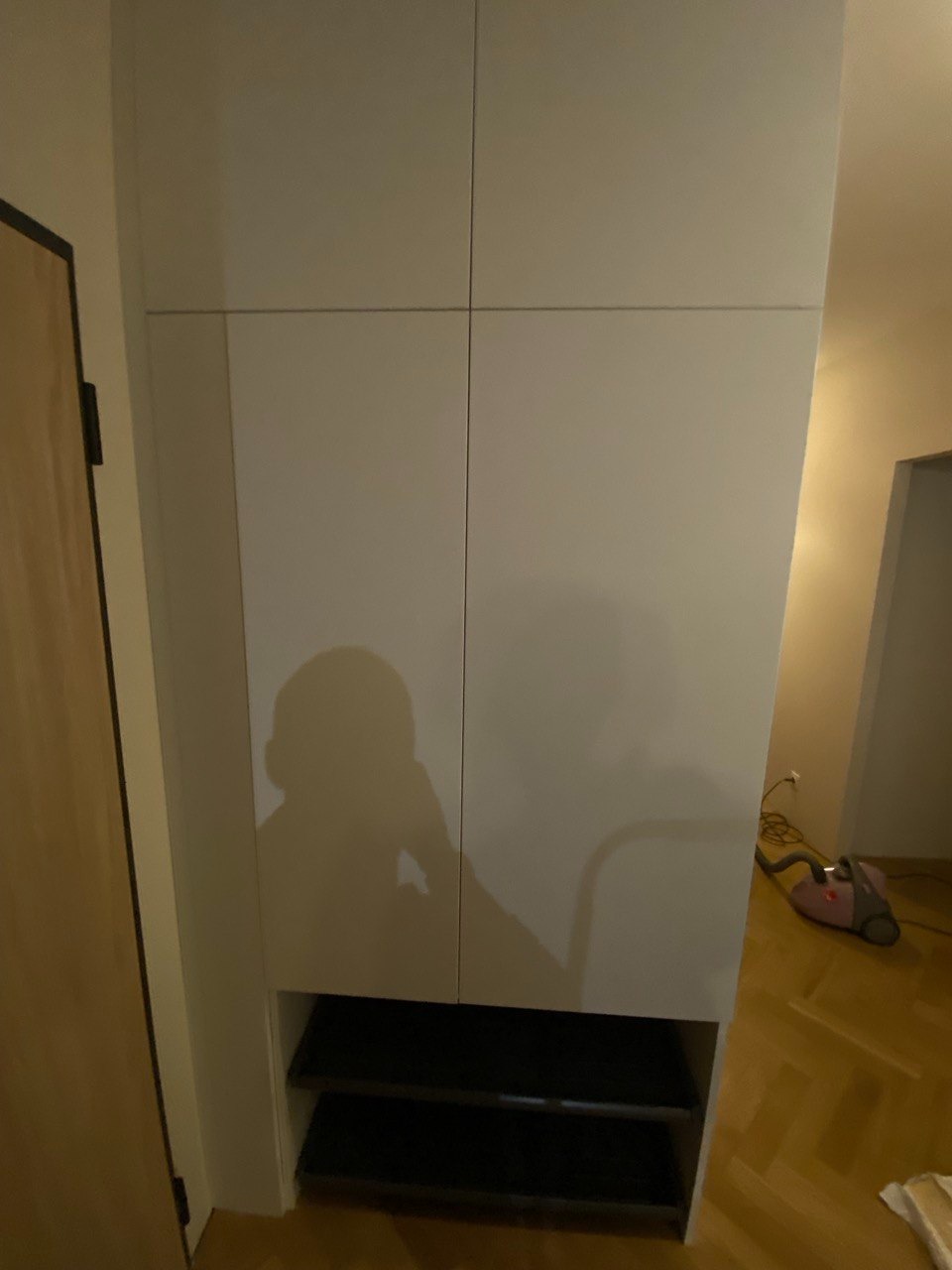 Biała szafa z frontami bez uchwytów i otwartą półką na dole, cień osoby widoczny na froncie, odkurzacz w tle.