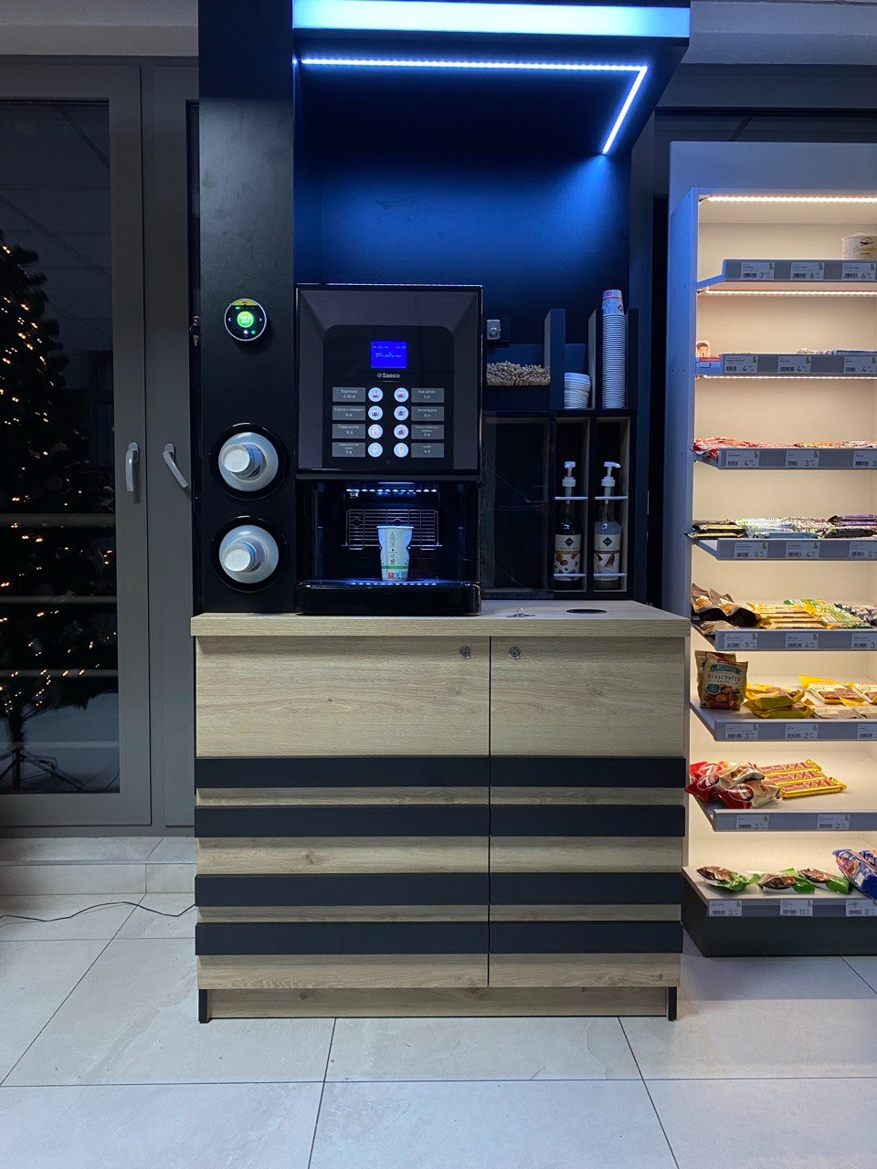 Automatyczny ekspres do kawy Saeco w zabudowie z szafkami w kolorze drewna i czarnymi pasami, obok regał z przekąskami, całość oświetlona niebieskim światłem LED.