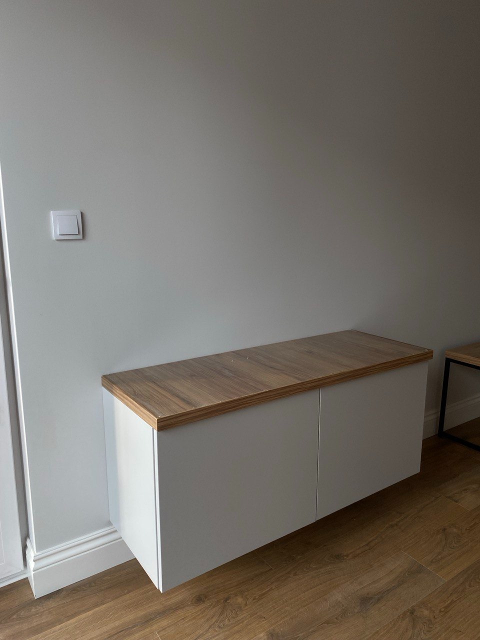 Biała szafka wisząca z blatem w kolorze drewna, zamontowana pod ścianą z włącznikiem światła, widoczna na tle drewnianej podłogi.