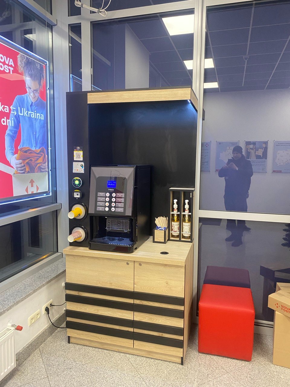 Automat do kawy w zabudowie meblowej z drewnianymi frontami i czarnymi akcentami, obok czerwony i fioletowy puf, widoczne okno z plakatem i odbijająca się sylwetka osoby.