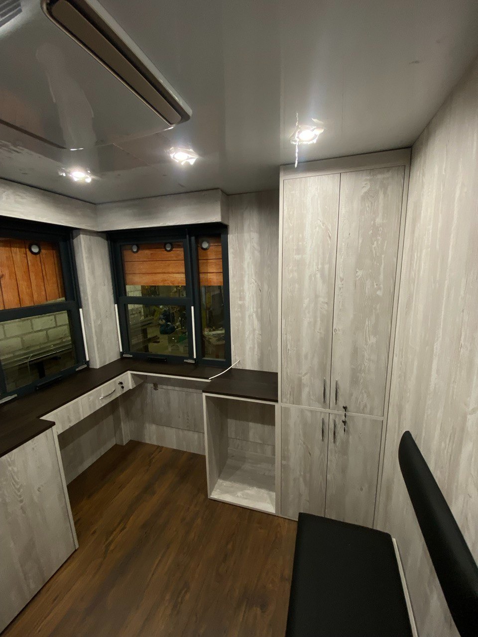 Wnętrze z zabudową meblową w odcieniach szarości i brązu: szafa zamykana na klucz, biurko z drewnianym blatem pod oknem z żaluzjami, drewniana podłoga.