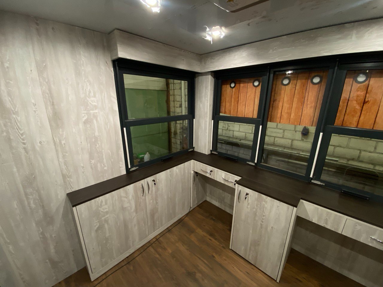 Szafki pod oknami z blatem w kolorze ciemnego brązu i frontami w odcieniu jasnego drewna, zamontowane w pomieszczeniu z oknami z ciemnymi ramami.