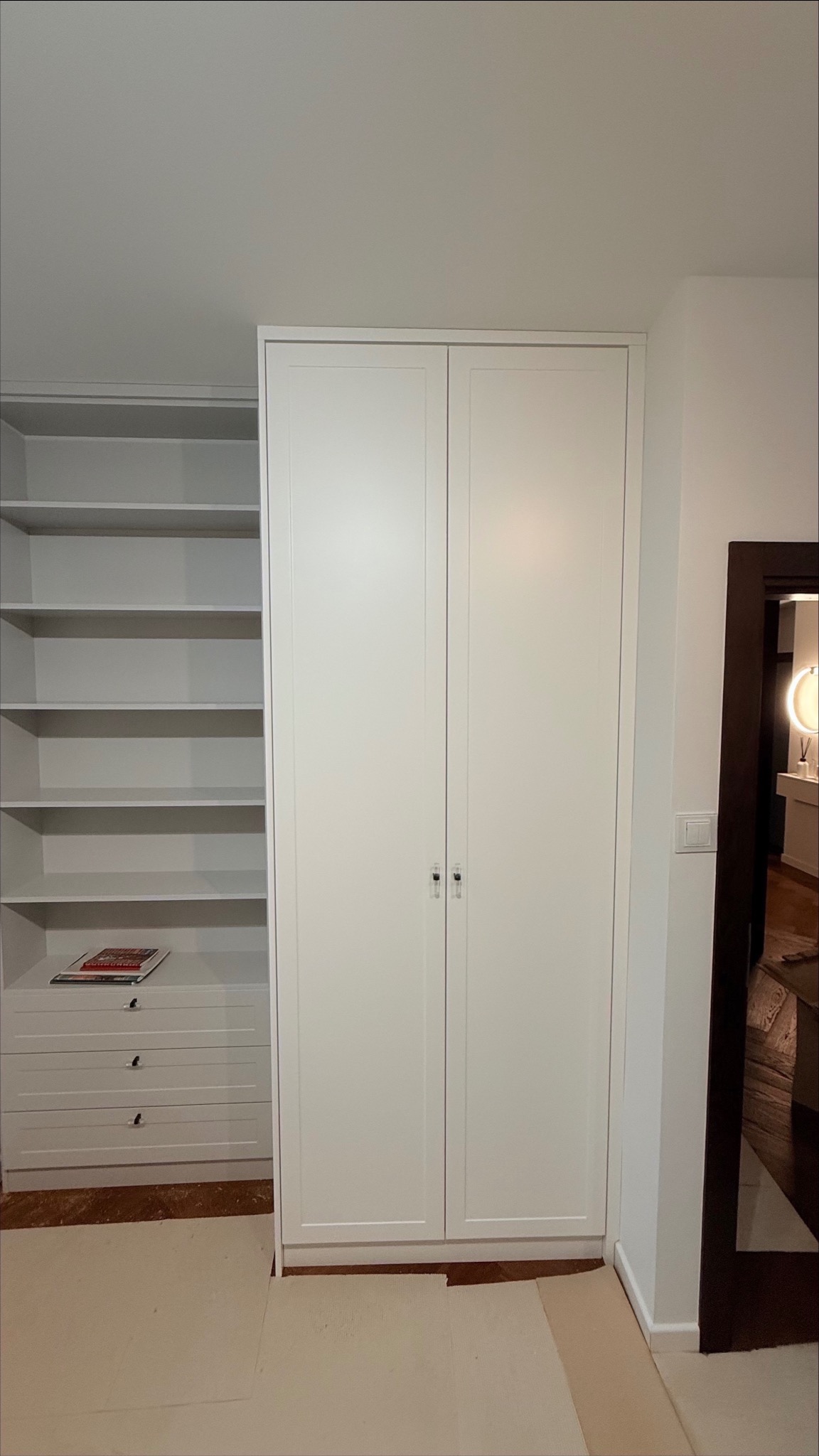 Biała szafa wnękowa z dwoma drzwiami i regał z półkami, widoczne w jasnym wnętrzu. Minimalistyczny design, proste uchwyty, subtelne oświetlenie.