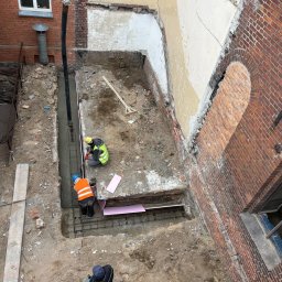 RENEWAL CONSTRUCTIONS SPÓŁKA Z OGRANICZONĄ ODPOWIEDZIALNOŚCIĄ - Wysoki kąt: robotnicy w kaskach przy wylewaniu fundamentów w wykopie obok ceglanej ściany z oknem i resztkami tynku. Pompa do betonu w rogu. Widoczne zbrojenia.