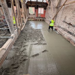 RENEWAL CONSTRUCTIONS SPÓŁKA Z OGRANICZONĄ ODPOWIEDZIALNOŚCIĄ - Wylewka podłogowa w remontowanym poddaszu kamienicy. Część powierzchni zalana, pracownik wyrównuje beton listwą wibracyjną. Widoczne stare cegły i drewniana konstrukcja dachu.