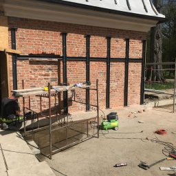 RENEWAL CONSTRUCTIONS SPÓŁKA Z OGRANICZONĄ ODPOWIEDZIALNOŚCIĄ - Wzmocnienie muru z czerwonej cegły stalowymi profilami. Widoczny fragment budynku z zabezpieczonym dachem i pracownik wykonujący prace montażowe przy rusztowaniu.