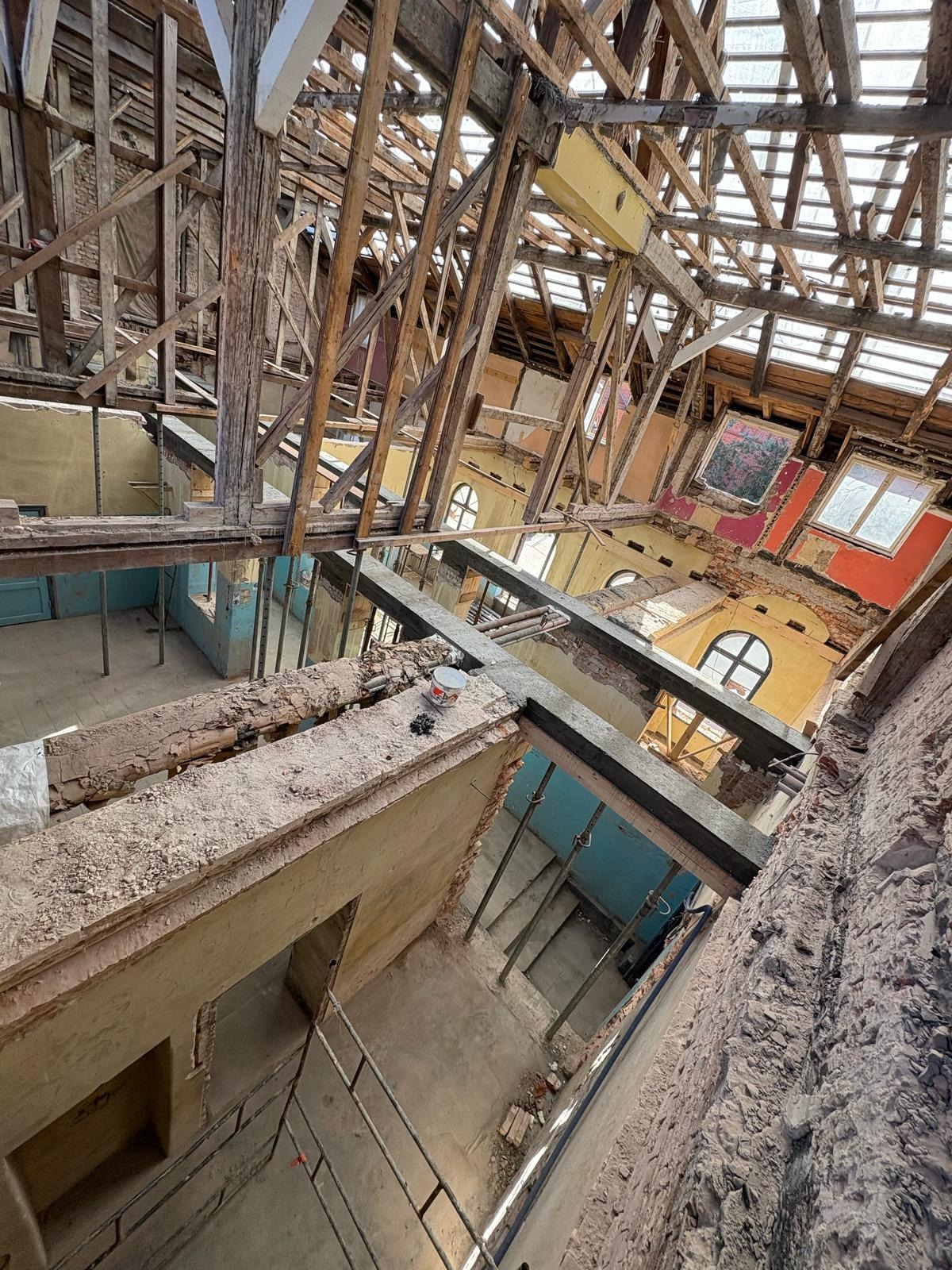 Wnętrze kamienicy w trakcie remontu: odsłonięta więźba dachowa, ściany z fragmentami farb, podpory stropowe i rusztowania w perspektywie z góry.