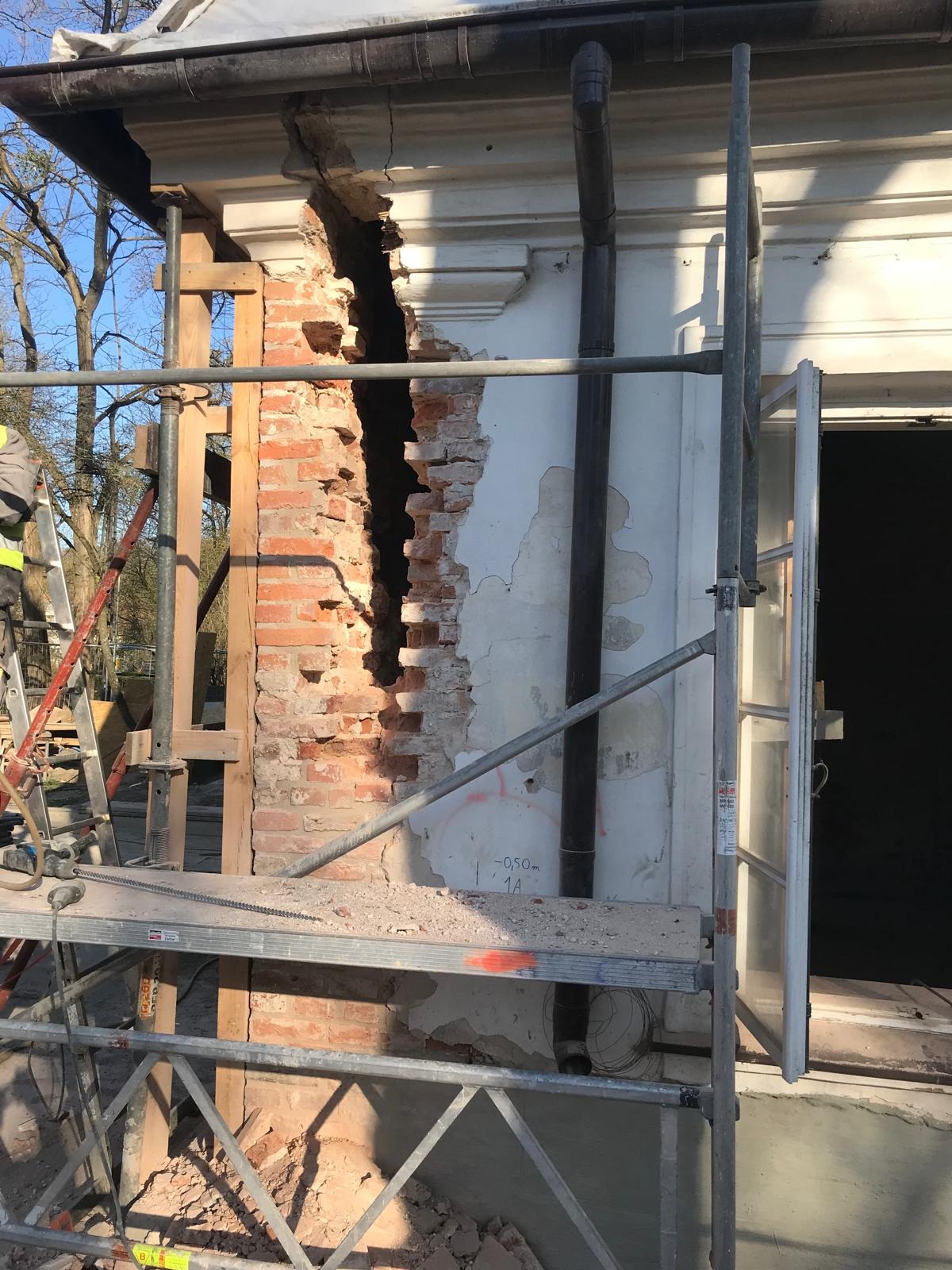 Uszkodzona ściana budynku z odsłoniętą cegłą, zabezpieczona rusztowaniem i drewnianą konstrukcją, widoczne ślady napraw i renowacji.