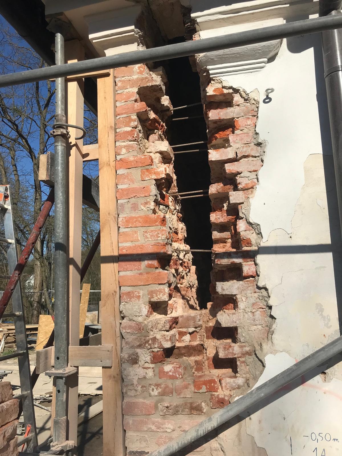 Fragment uszkodzonej ściany z cegły z widocznym zbrojeniem, obok fragment białej ściany z ubytkiem tynku. Widoczne rusztowanie i drewniane elementy konstrukcyjne.