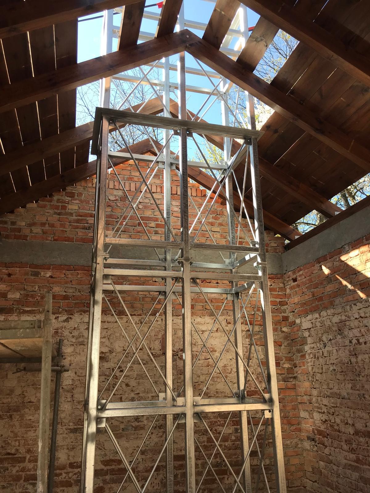 Wnętrze budynku w trakcie remontu. Metalowa konstrukcja, odsłonięta cegła, drewniane belki stropowe i widok na niebo przez otwór w dachu.