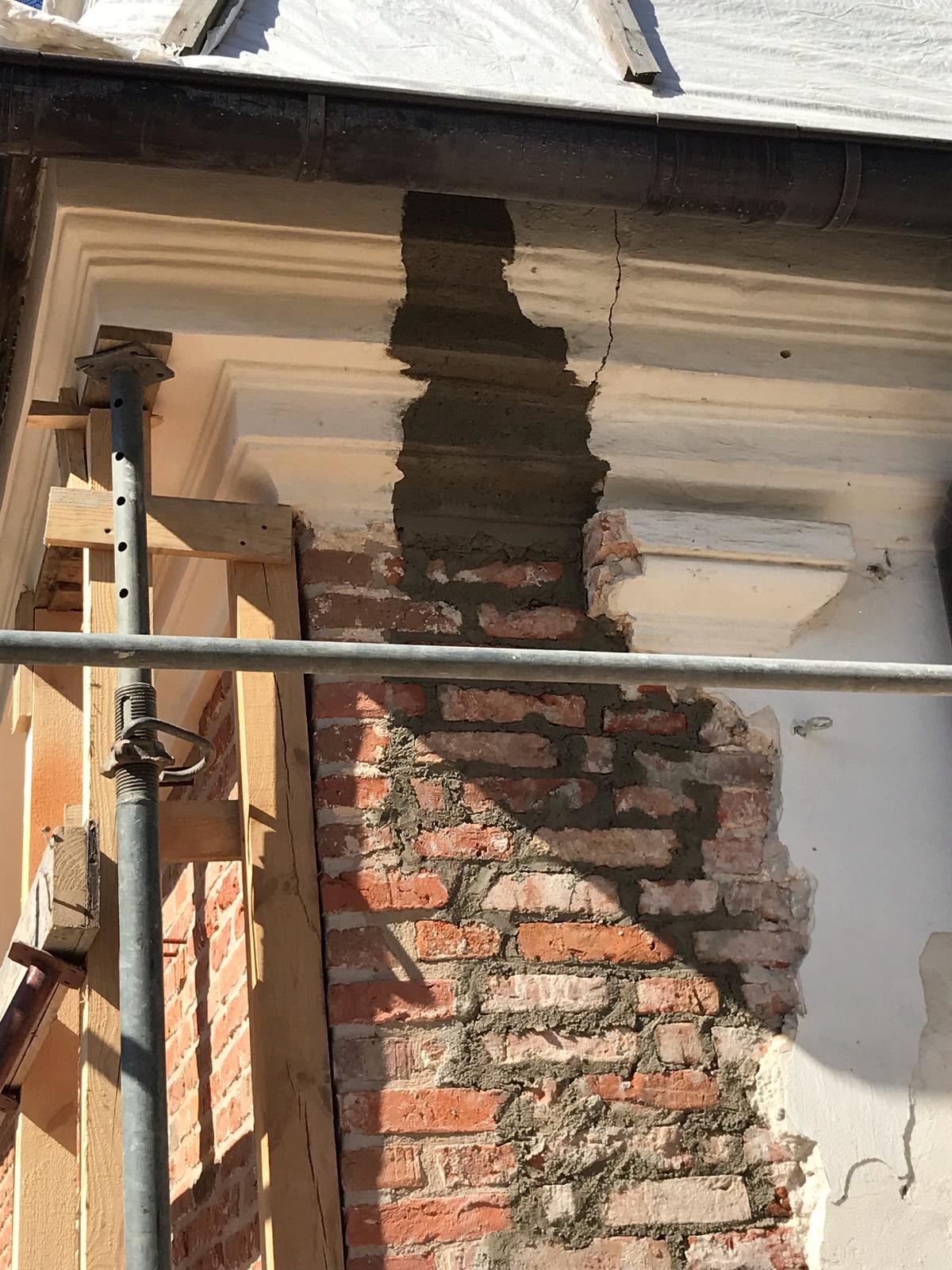 Fragment elewacji budynku w trakcie renowacji, z widocznym murem z czerwonej cegły i częściowo odnowionym, białym elementem dekoracyjnym. Widoczne rusztowanie.