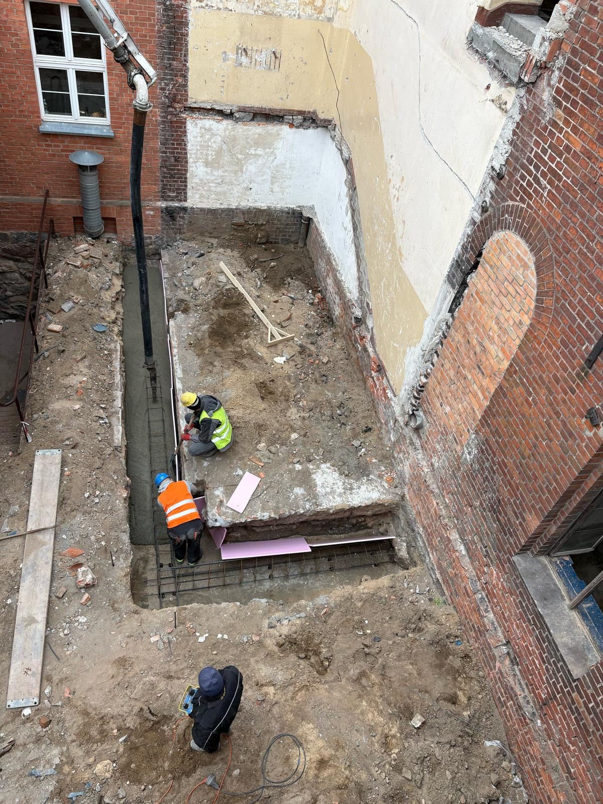 Wysoki kąt: robotnicy w kaskach przy wylewaniu fundamentów w wykopie obok ceglanej ściany z oknem i resztkami tynku. Pompa do betonu w rogu. Widoczne zbrojenia.