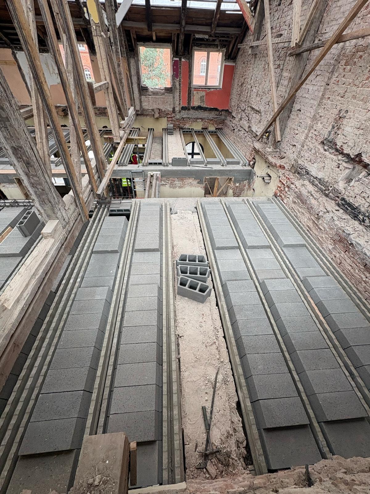 Wnętrze remontowanego budynku z widocznymi bloczkami betonowymi ułożonymi w rzędach, drewnianą konstrukcją dachu i fragmentami odsłoniętej cegły na ścianach.