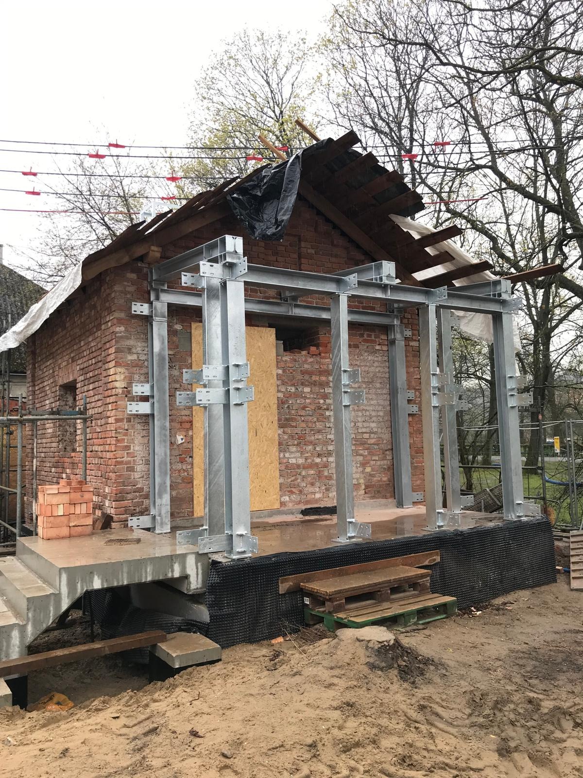 Ceglany budynek w trakcie rozbudowy z widocznymi stalowymi elementami konstrukcyjnymi. Częściowo odsłonięty dach i fundamenty. Teren budowy.