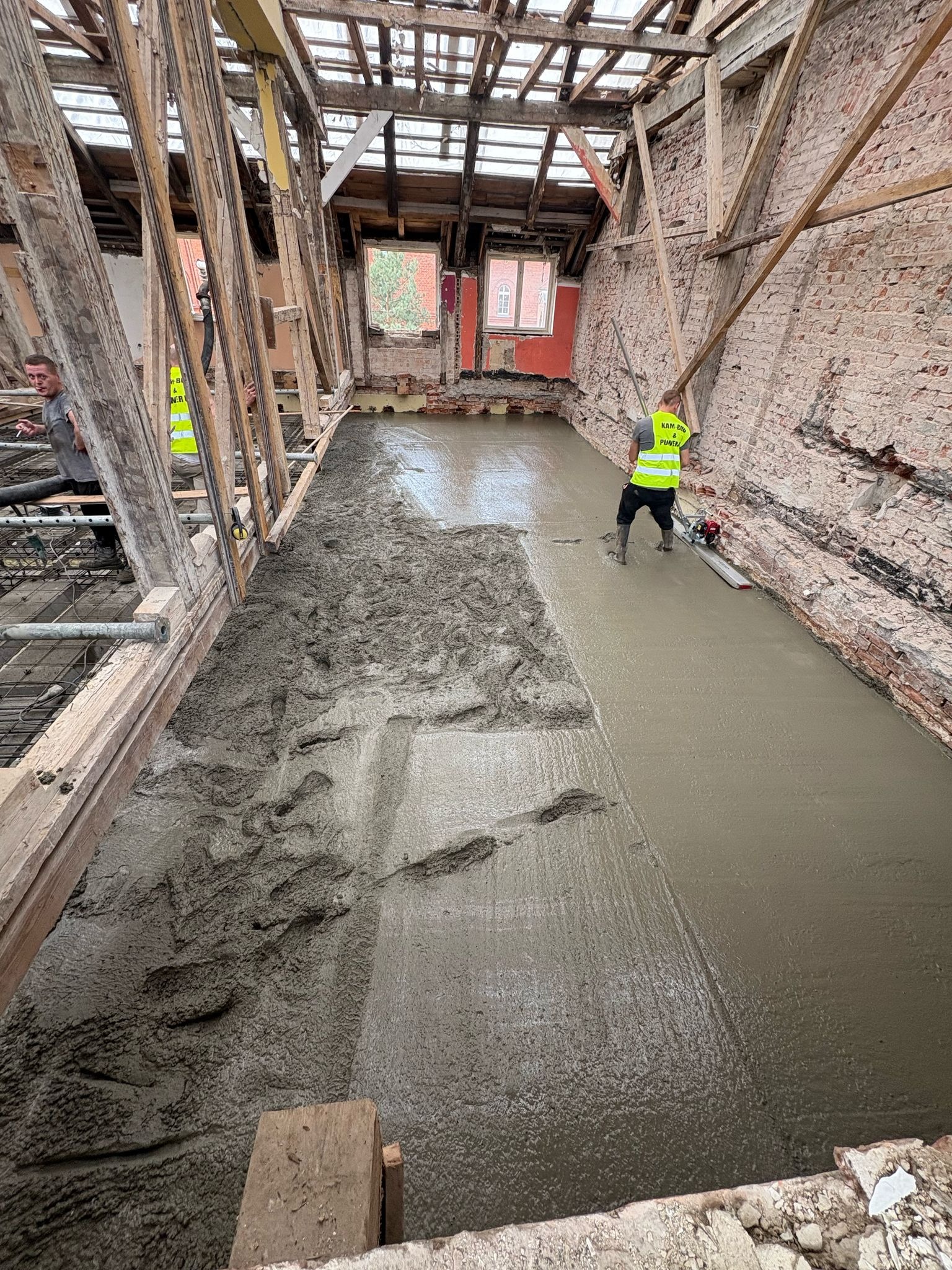 Wylewka podłogowa w remontowanym poddaszu kamienicy. Część powierzchni zalana, pracownik wyrównuje beton listwą wibracyjną. Widoczne stare cegły i drewniana konstrukcja dachu.
