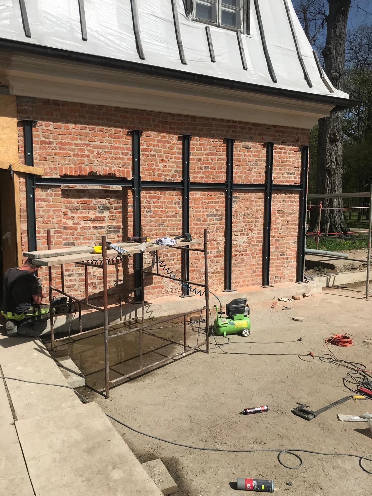 Wzmocnienie muru z czerwonej cegły stalowymi profilami. Widoczny fragment budynku z zabezpieczonym dachem i pracownik wykonujący prace montażowe przy rusztowaniu.