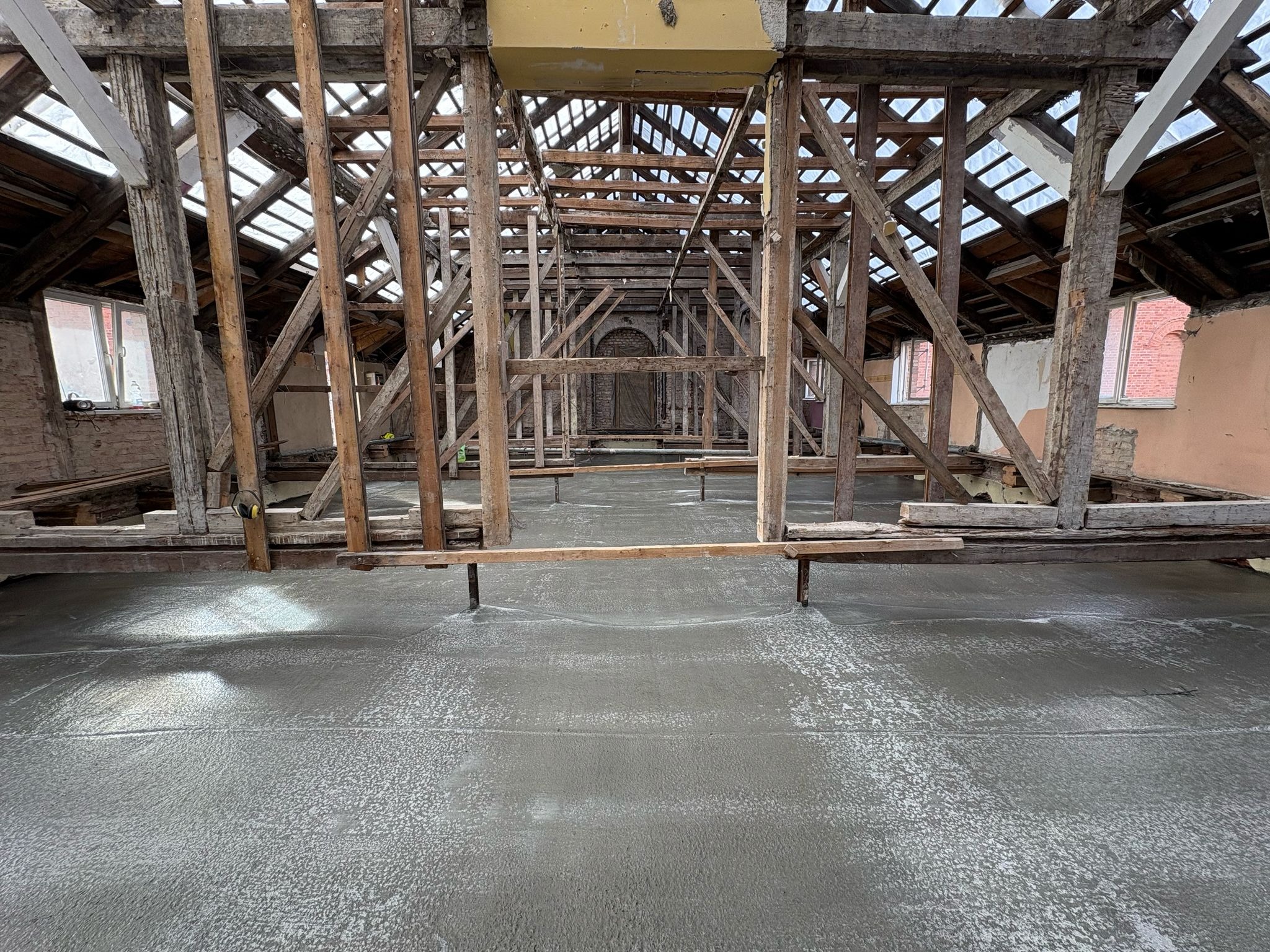 Wnętrze remontowanego poddasza z odsłoniętą konstrukcją drewnianą i świeżo wylaną, jeszcze mokrą, betonową posadzką. Widoczne okna i fragmenty muru.
