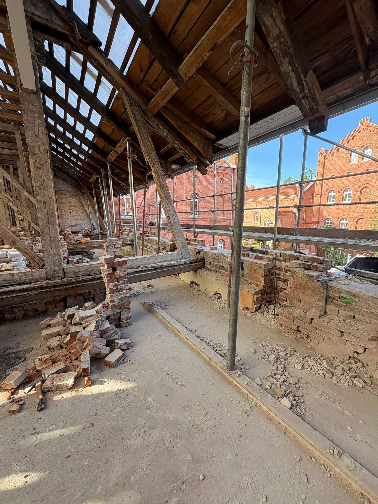 Wnętrze poddasza w trakcie remontu: odsłonięta drewniana konstrukcja dachu, fragmenty ceglanej ściany, rusztowania i porozrzucane cegły. Widok na ceglany budynek z zewnątrz.
