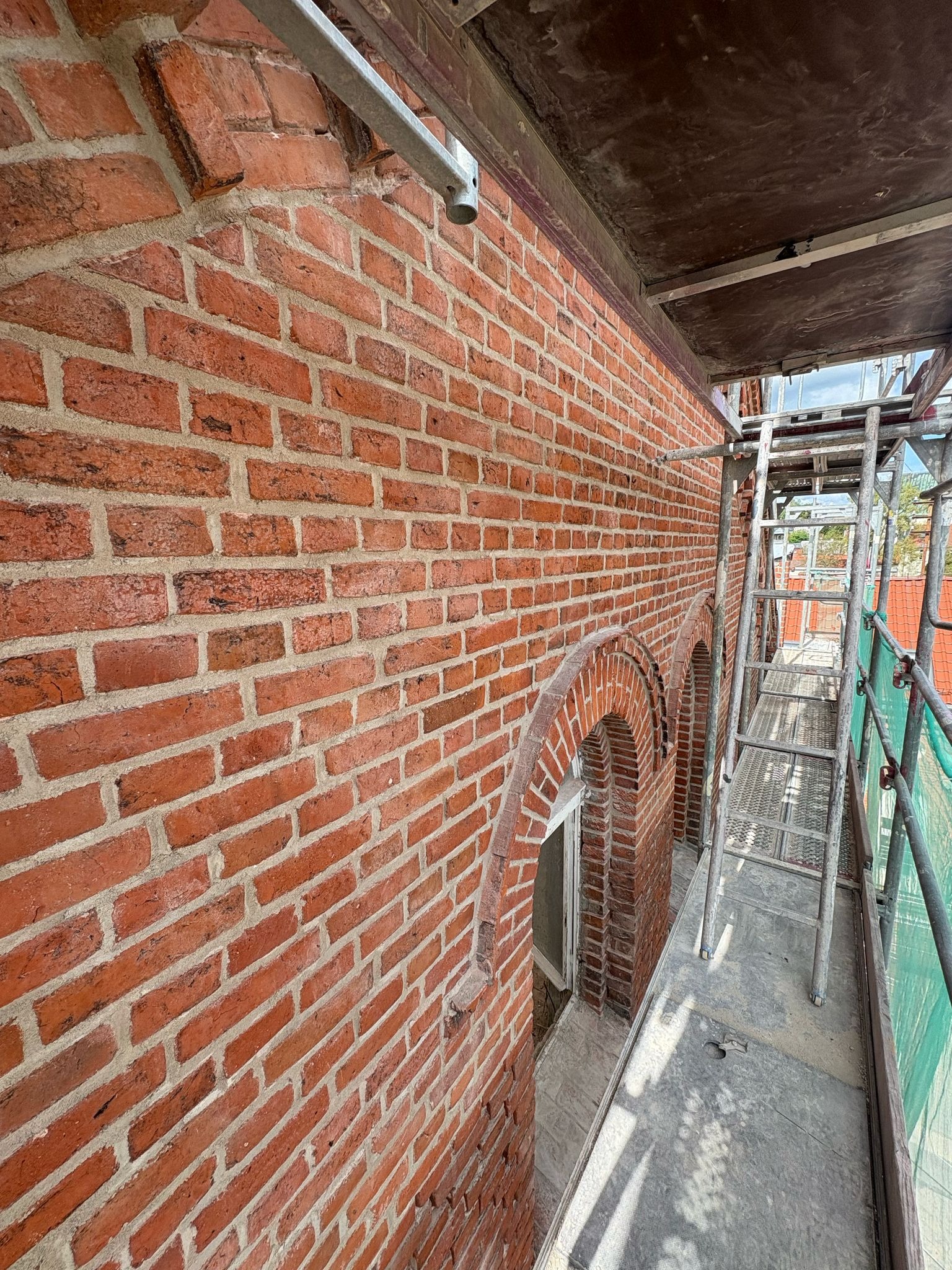 Ceglana ściana budynku z elementami łukowymi, widoczna z poziomu rusztowania. Detale murarskie i struktura cegieł w bliskim ujęciu.