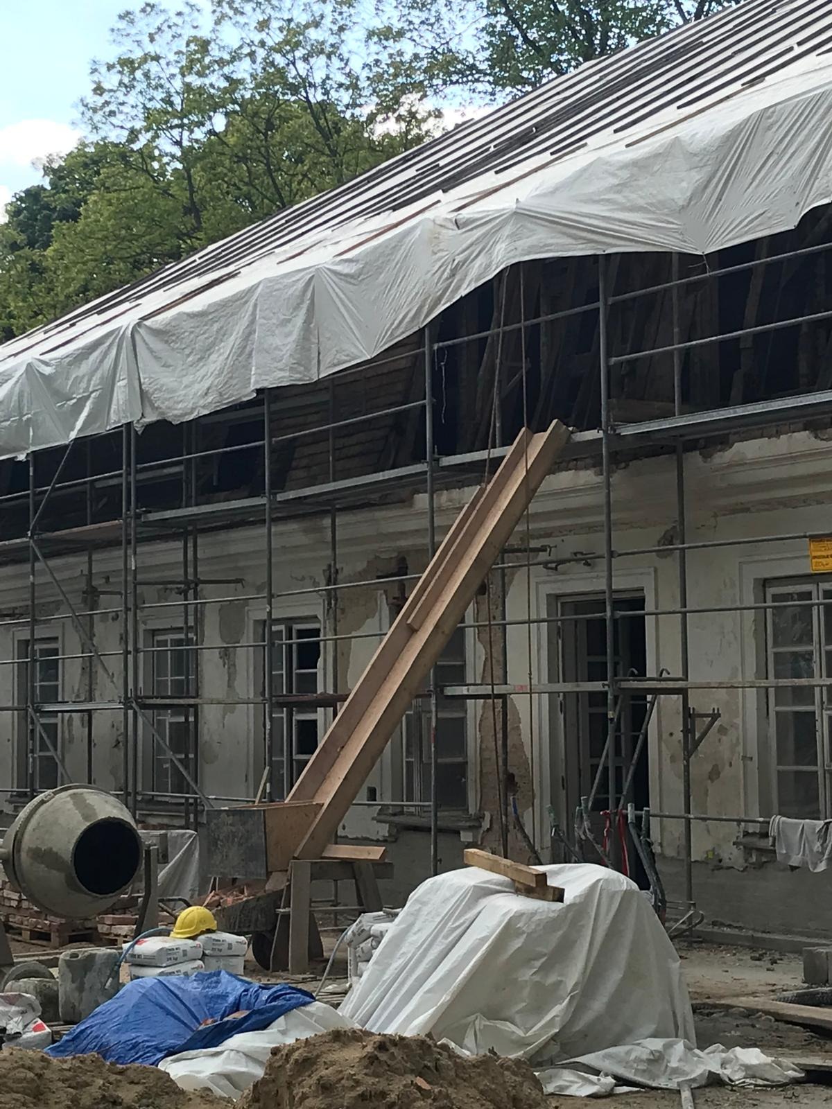 Remont elewacji kamienicy ze rusztowaniem i zsypem. Fragment odkrytego dachu pod plandeką. Widoczny mikser budowlany i teren budowy.