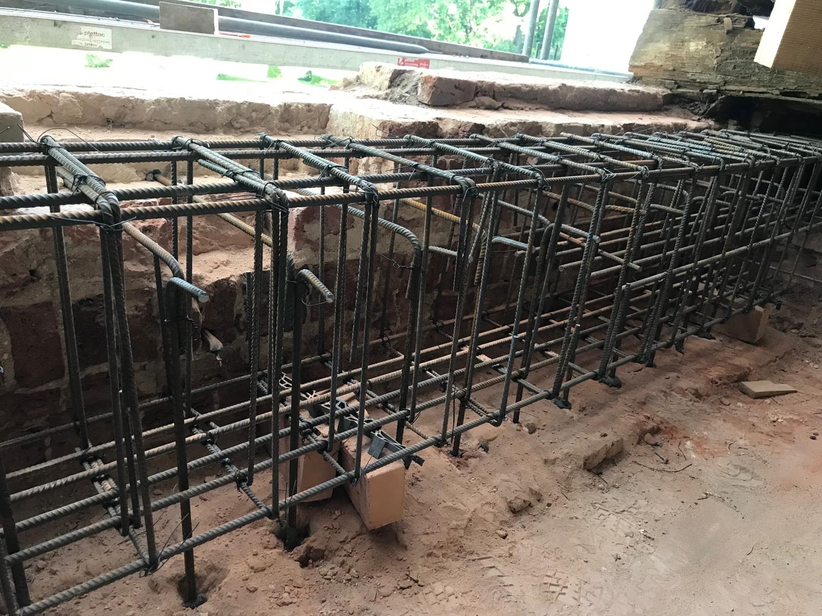Zbrojenie fundamentu z prętów stalowych, przygotowane do zalania betonem, widoczne ceglane elementy konstrukcyjne w tle, grunt piaszczysty.