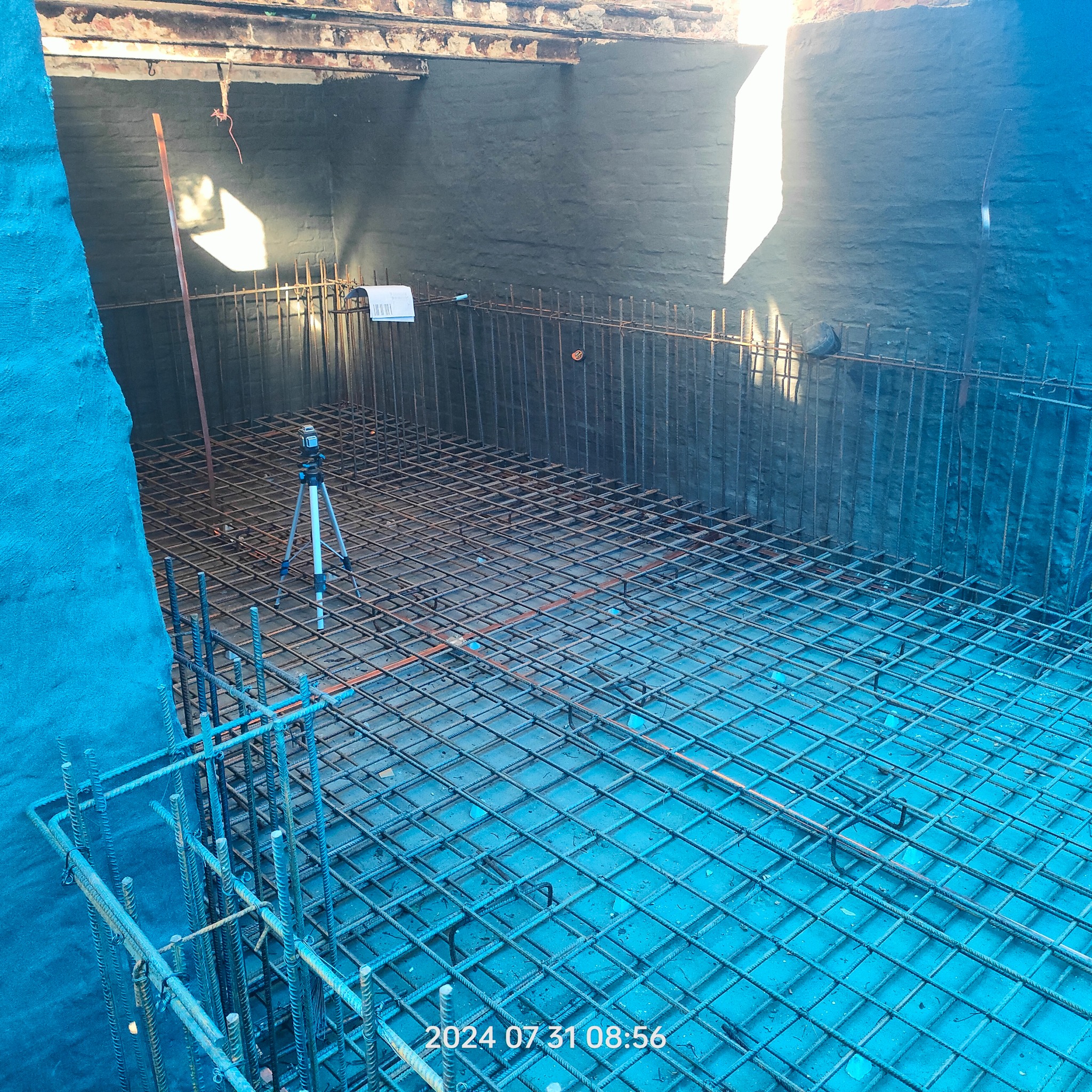 Zbrojenie fundamentu z użyciem stalowych prętów, tworzących siatkę. Na pierwszym planie pionowe pręty. W tle ściana z cegieł i laserowy niwelator na statywie.