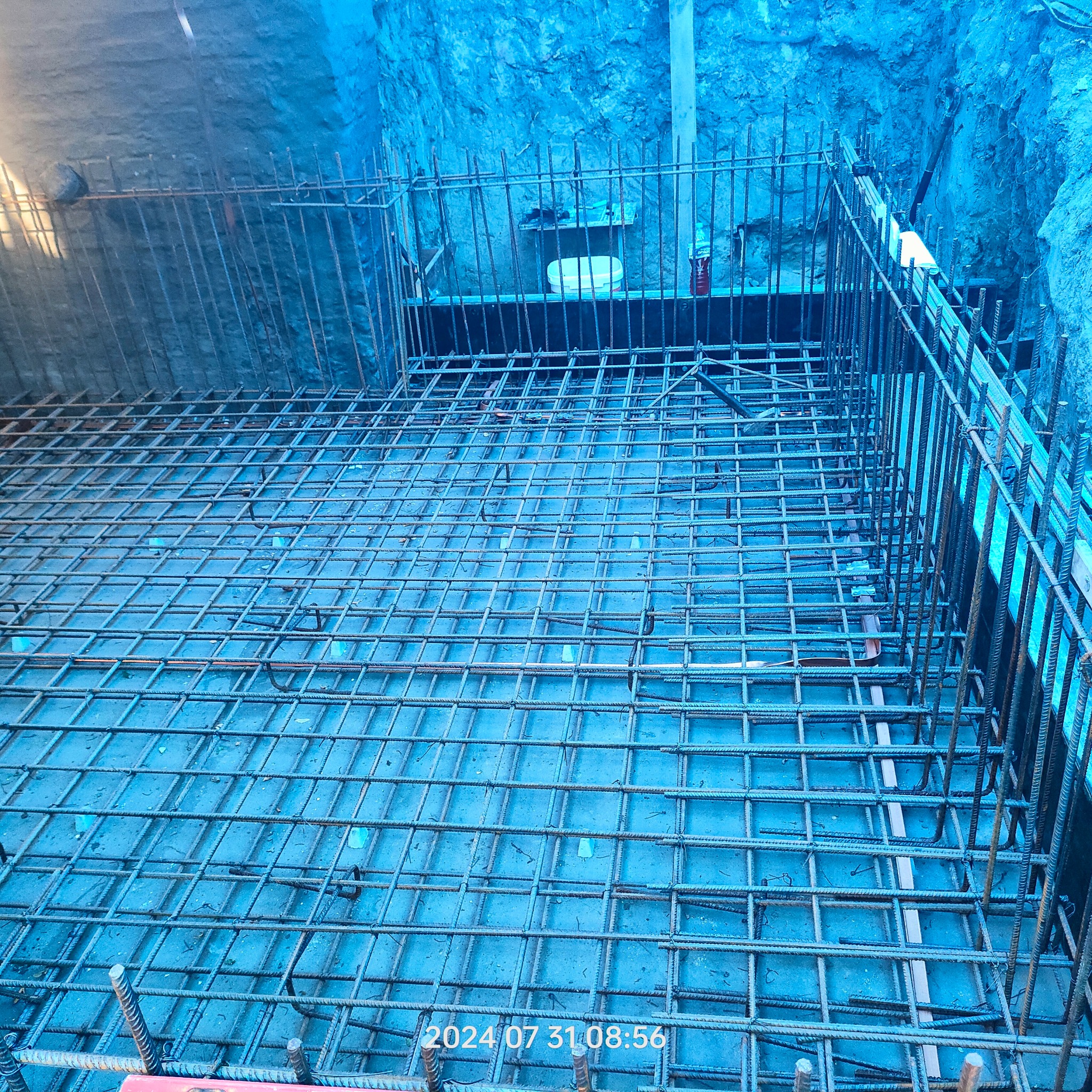 Zbrojenie fundamentów w wykopie, widoczne stalowe pręty i siatka zbrojeniowa. Ściany wykopu zabezpieczone folią. Data i godzina na zdjęciu.