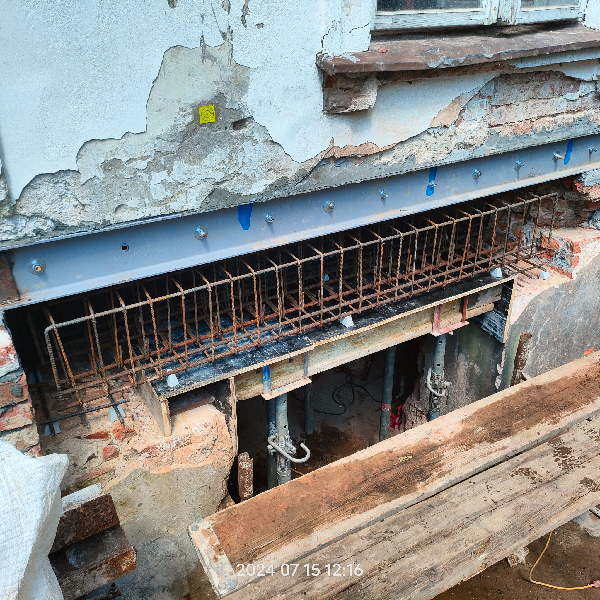 Wzmocnienie fundamentów starego domu z odsłoniętą cegłą. Stalowe belki i zbrojenie podtrzymują konstrukcję. Widoczny fragment okna i zniszczony tynk.