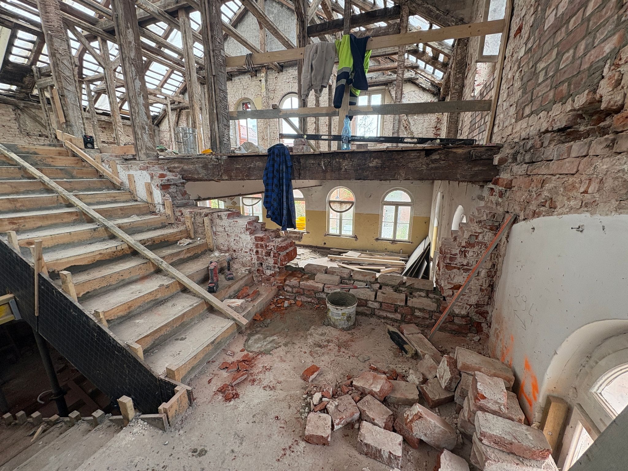 Wnętrze remontowanego budynku z odsłoniętą konstrukcją drewnianą dachu, schodami w trakcie budowy i fragmentami muru z czerwonej cegły. Widoczne narzędzia i materiały budowlane.