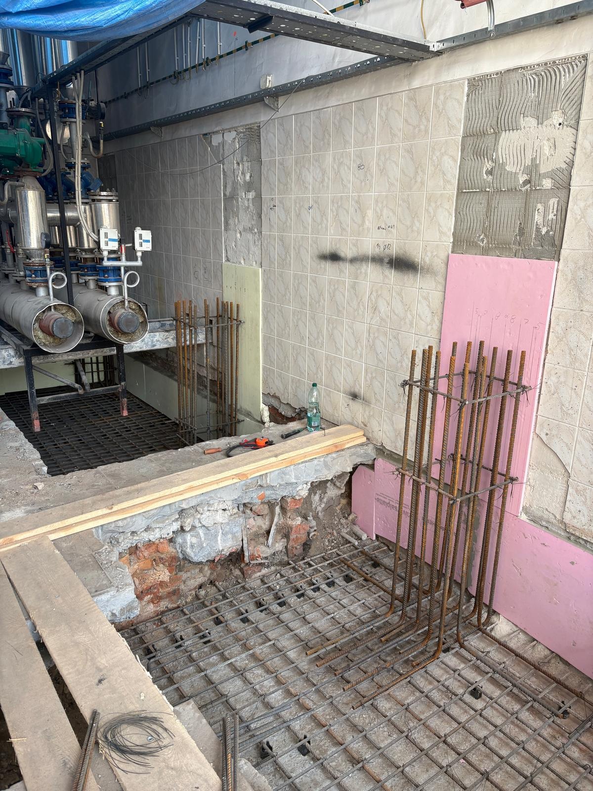 Zbrojenie pod wylewkę w pomieszczeniu z częściowo usuniętymi płytkami. Widoczne narzędzia, butelka wody oraz instalacja rurowa na ścianie.