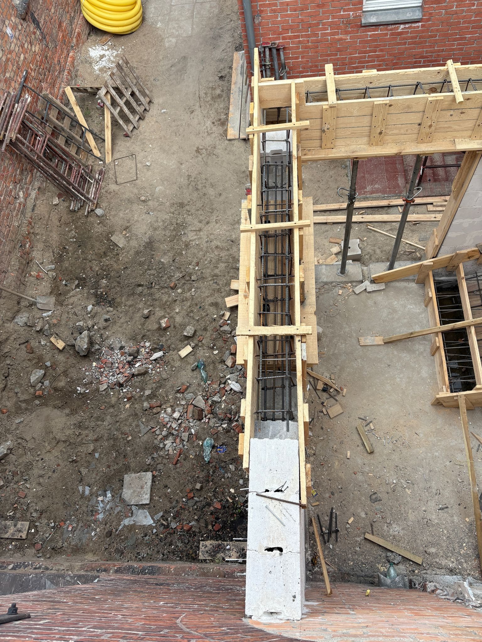 Wysoki kąt: zbrojenie pionowe w szalunku na placu budowy, widoczne ceglane ściany i gruz. Żółte rury w tle. Etap prac fundamentowych.