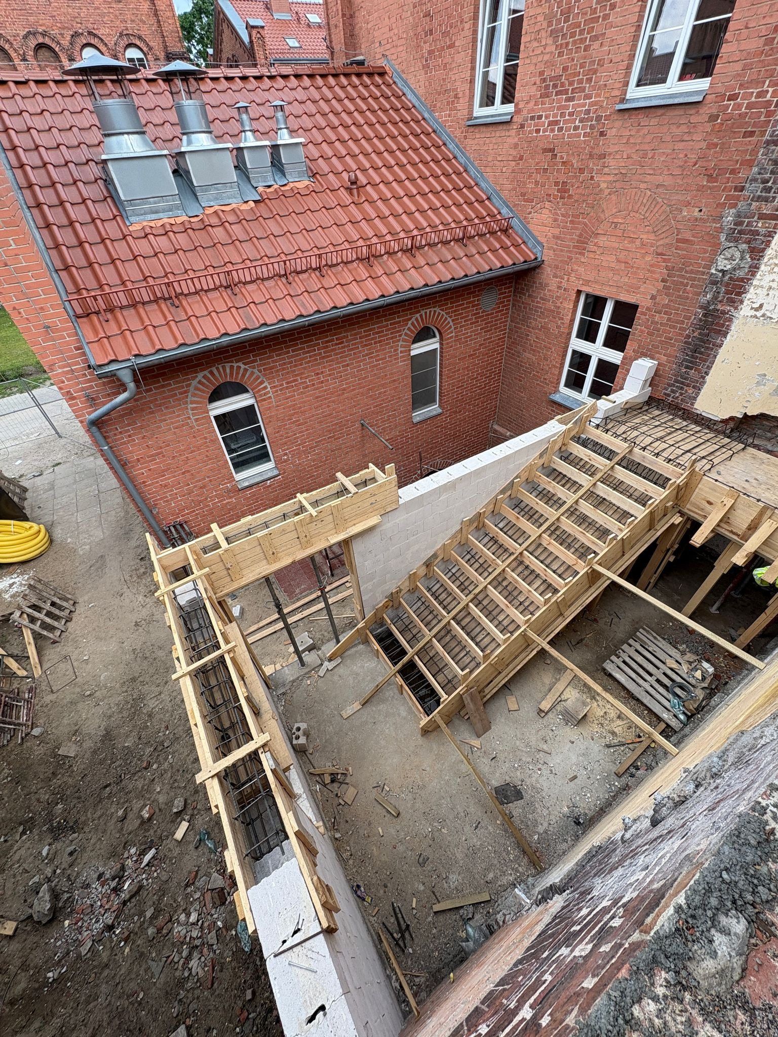 Budowa schodów w trakcie realizacji w Warszawie. Widoczne szalunki i zbrojenia, ceglany budynek w tle. Kąt ujęcia z góry, ukazujący postęp prac.