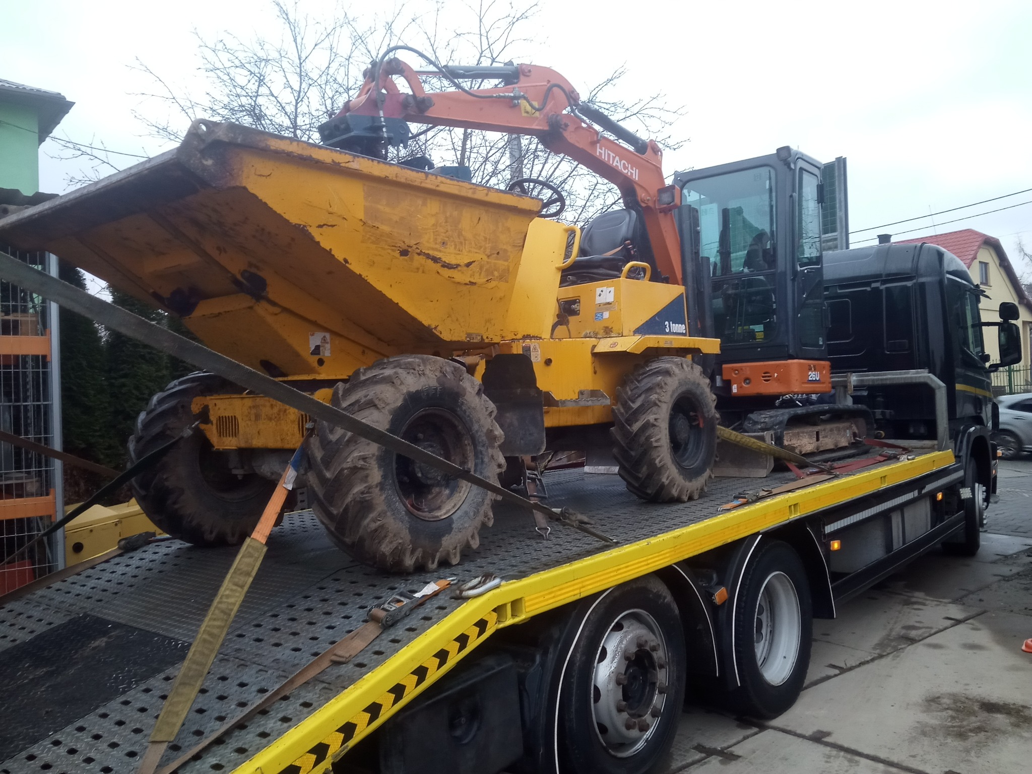 Żółta wywrotka Hitachi 3-tonowa z koparką na platformie lawety, zabezpieczona pasami transportowymi. Widoczne brudne opony i elementy hydrauliczne.