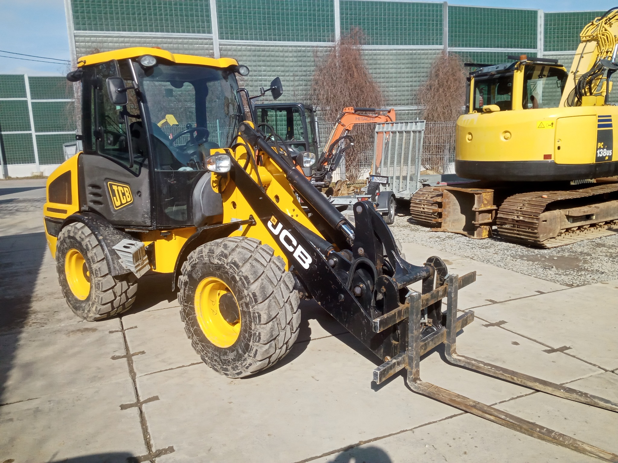 Żółto-czarna ładowarka JCB z widłami na tle betonowego placu i ekranów akustycznych, obok koparka Komatsu i mniejsza koparka na przyczepie.