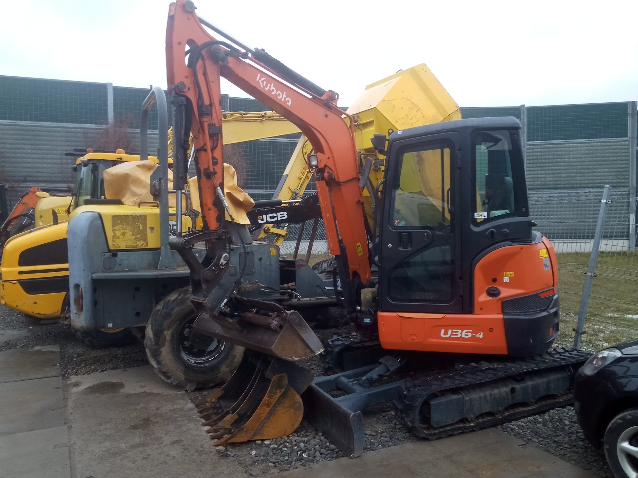 Pomarańczowa minikoparka Kubota U36-4 na żwirowym podłożu, z widocznymi łyżkami koparkowymi, w tle inne maszyny budowlane i ekran akustyczny.