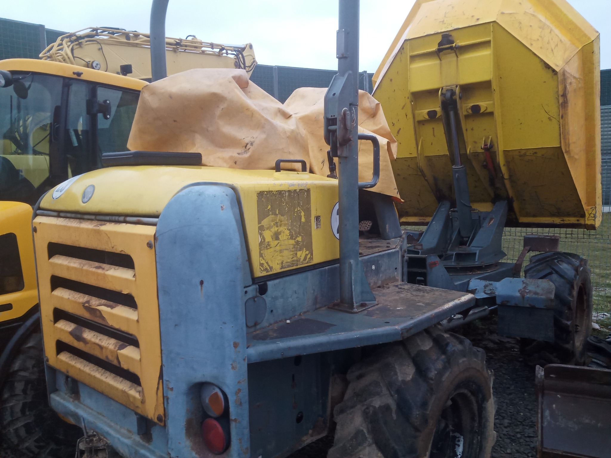 Żółto-szary wywrotka budowlana z podniesioną skrzynią, widoczne ślady użytkowania i zarysowania, zaparkowana na żwirowym terenie, obok żółta koparka z zakrytym brezentem siedzeniem.