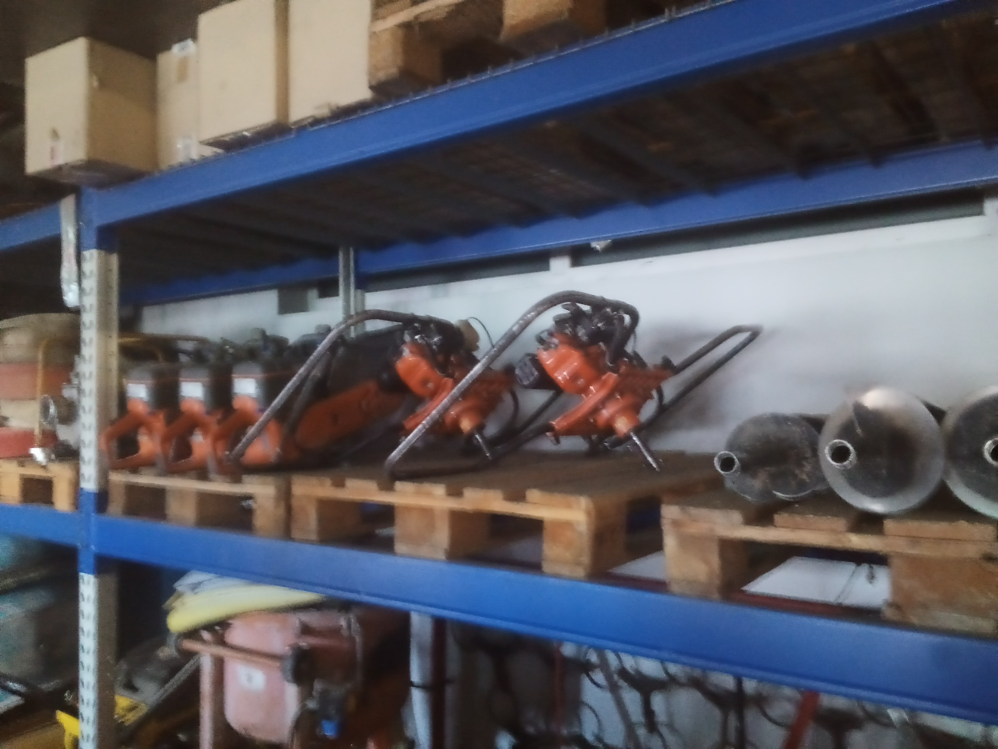 Magazynowe ujęcie kilku zagęszczarek w kolorze pomarańczowym, ustawionych na drewnianych paletach na metalowym regale z niebieskimi poprzeczkami.