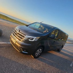 Twój-BUS - Czarny bus Renault Trafic zaparkowany na asfalcie z widokiem na morze i zachodzące słońce, zarys żaglówki na horyzoncie.
