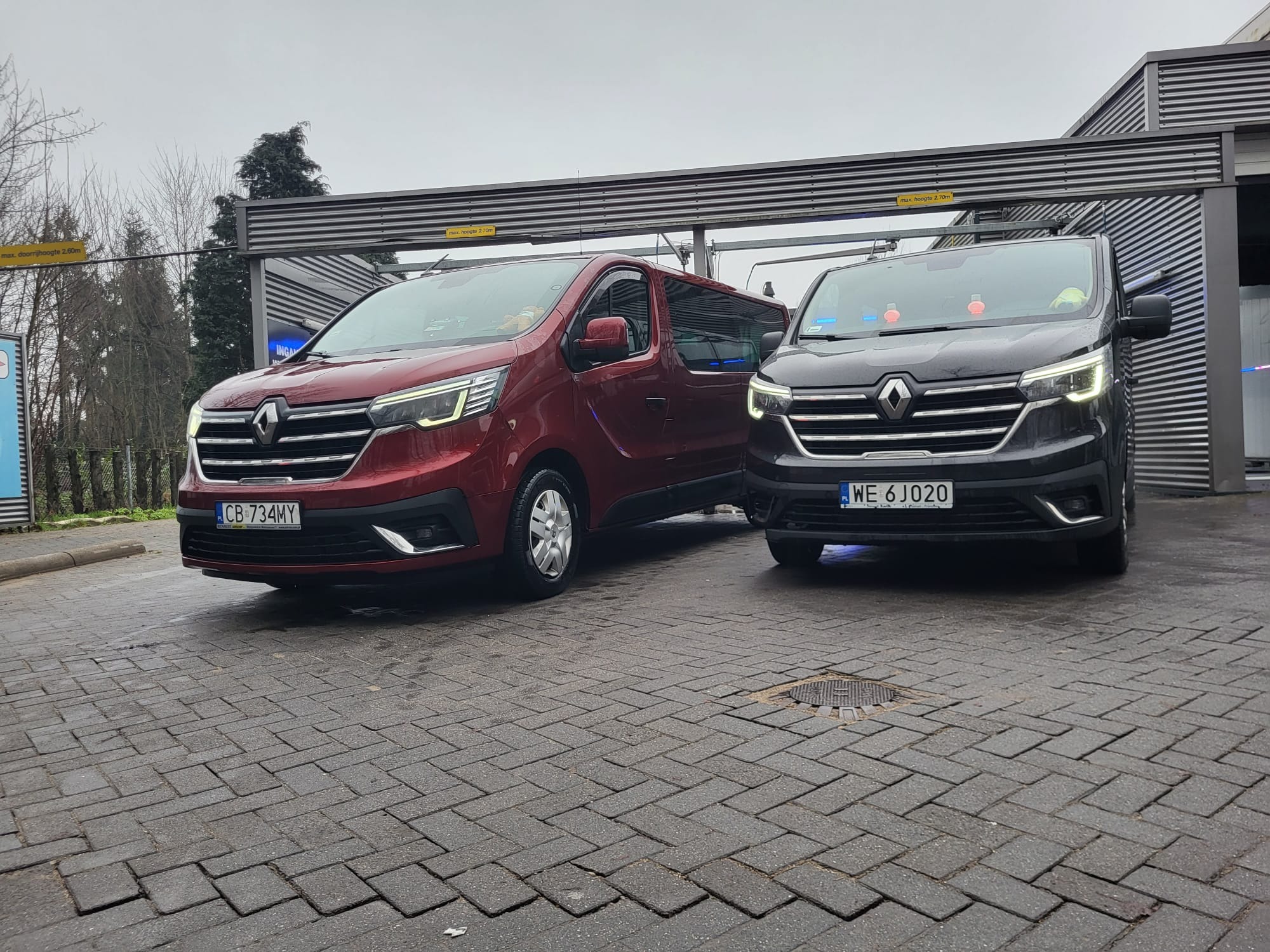 Dwa busy Renault Trafic: bordowy i szary, zaparkowane na mokrym bruku pod zadaszeniem. Widoczne tablice rejestracyjne CB 734MY i WE 6J020.